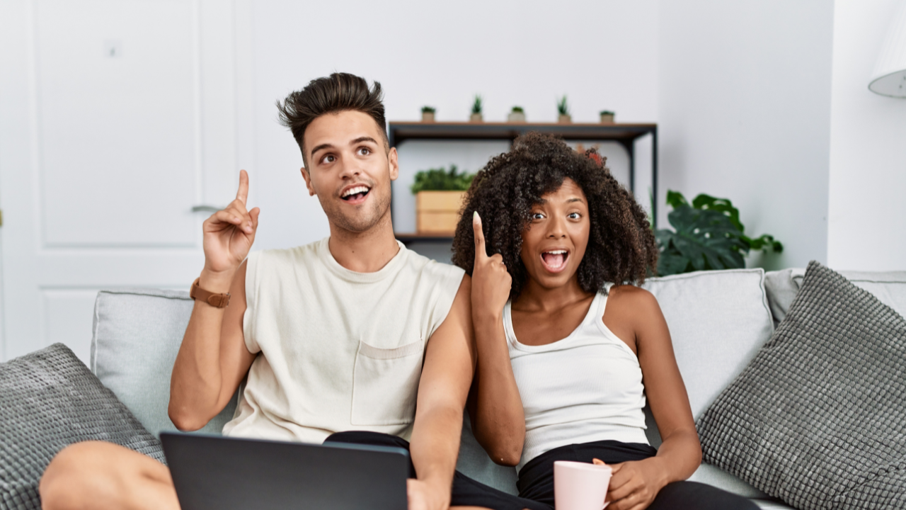 In the photo, a young couple is captured in a delightful moment of discovery. They are seated comfortably on a couch in a well lite living room 
setting.  Both individuals have their mouths open in an "aha!" expression, clearly excited about something they’ve just realized. The man and woman are pointing upwards, likely indicating a breakthrough or important idea they've come across. A laptop is open in front of them, suggesting that their moment of insight is related to something they found or figured out on the computer. The scene is filled with a sense of excitement and shared joy, highlighting their connection and enthusiasm.