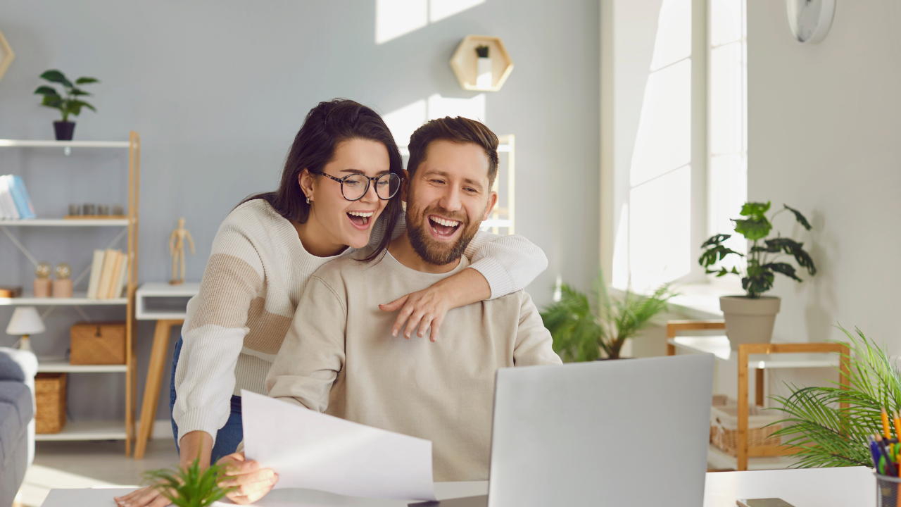 In the photo, a happy couple is sharing a joyful moment as they look at a computer screen together. The man has a broad smile on his face, clearly delighted by what he sees. The woman, equally joyous, has her arm lovingly draped around his neck, drawing them closer in a warm embrace. Their eyes are focused on the screen.  The scene exudes a sense of togetherness, comfort, and mutual support, encapsulating a beautiful moment of shared happiness and connection.